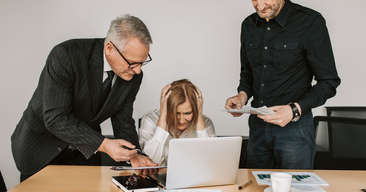 Three business professionals, including an elderly person, in a tense meeting around a laptop.