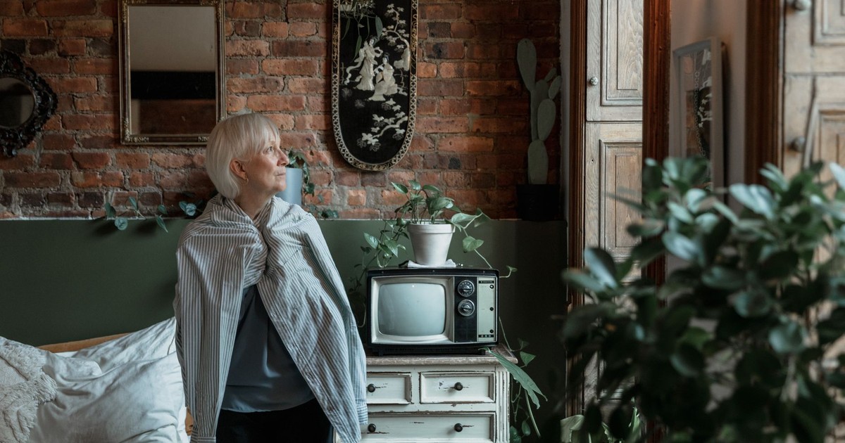 Elderly woman looks out window in cozy rustic room, surrounded by plants and vintage decor.