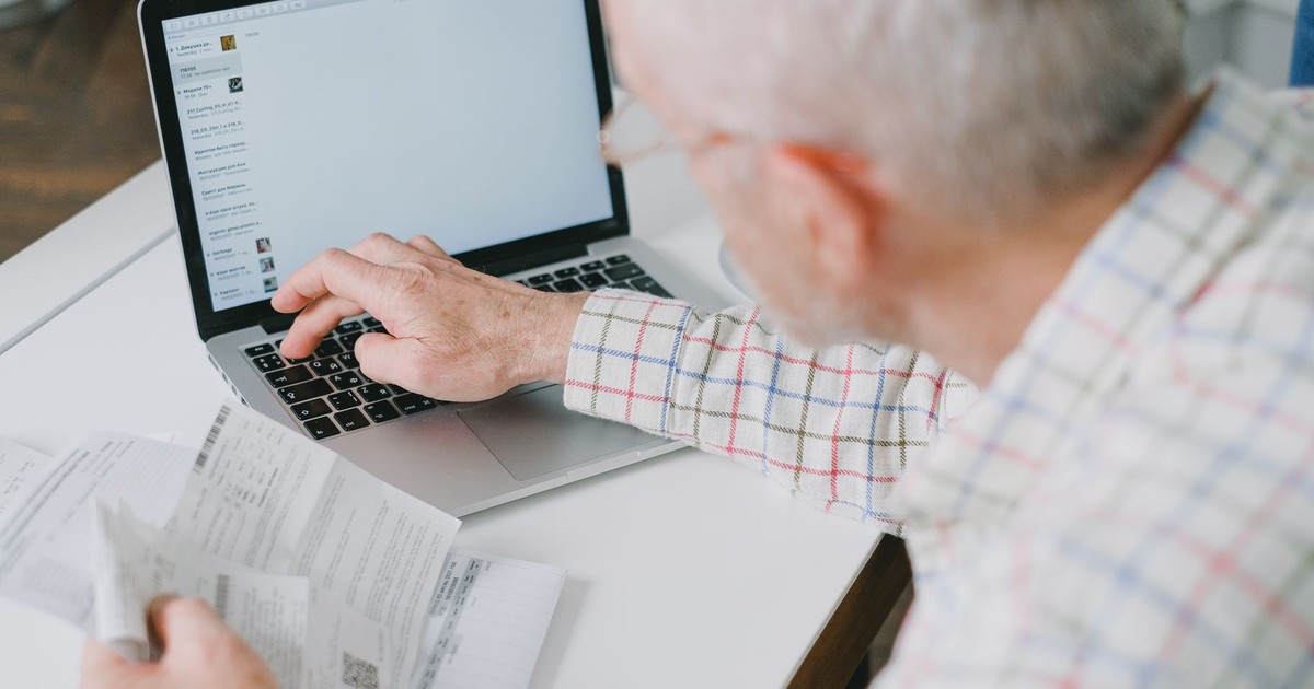 Elderly man at desk handling finances online with documents and laptop.