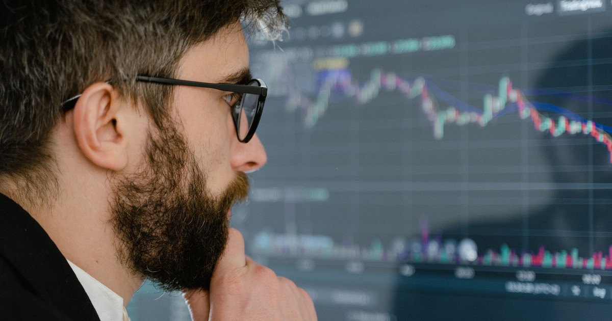 A focused man with glasses studies stock market graphs on a screen, pondering insights.