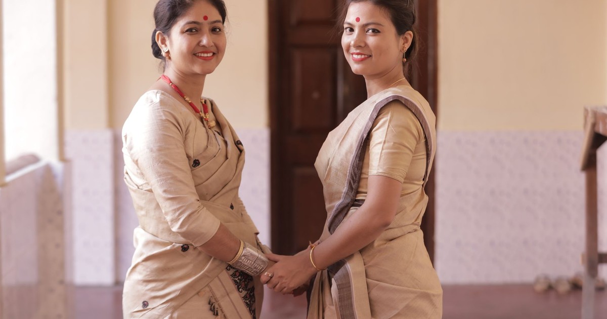 Two South Asian women in traditional sarees smiling indoors, reflecting Indian cultural attire.