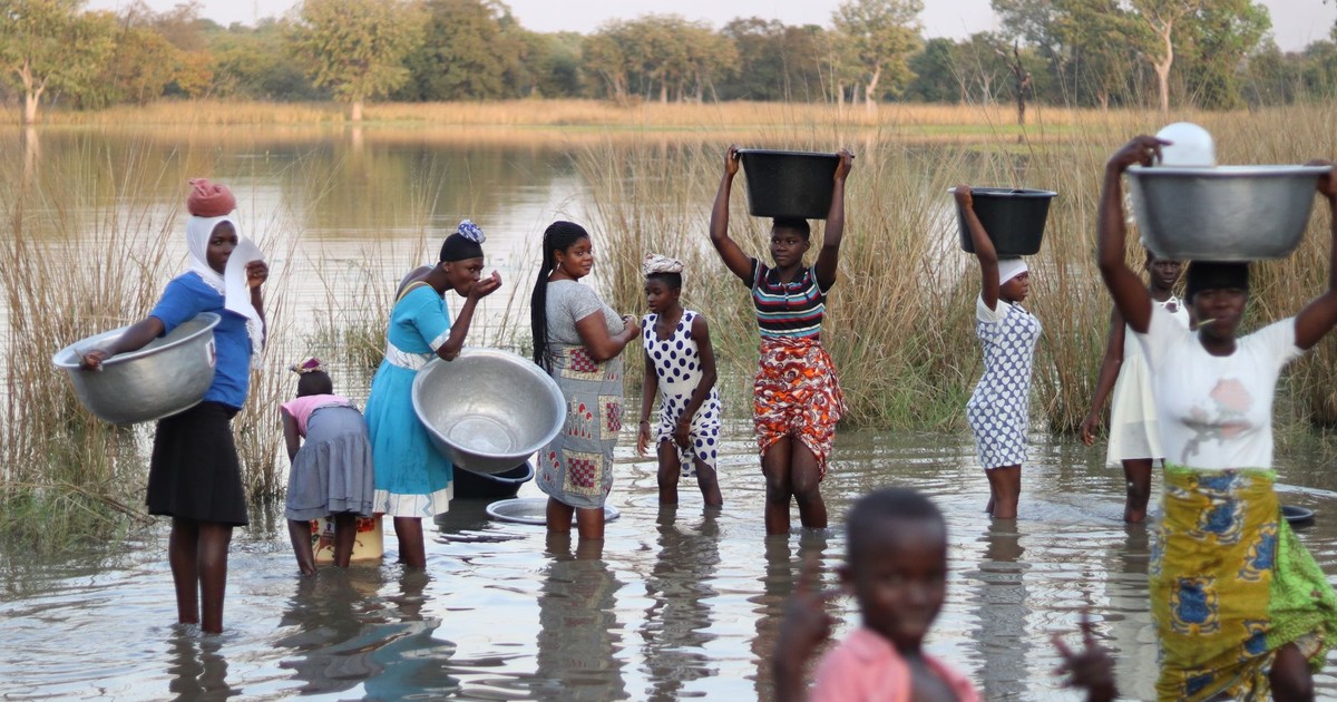 Group of women and children gathering water from a river using metal bowls.
