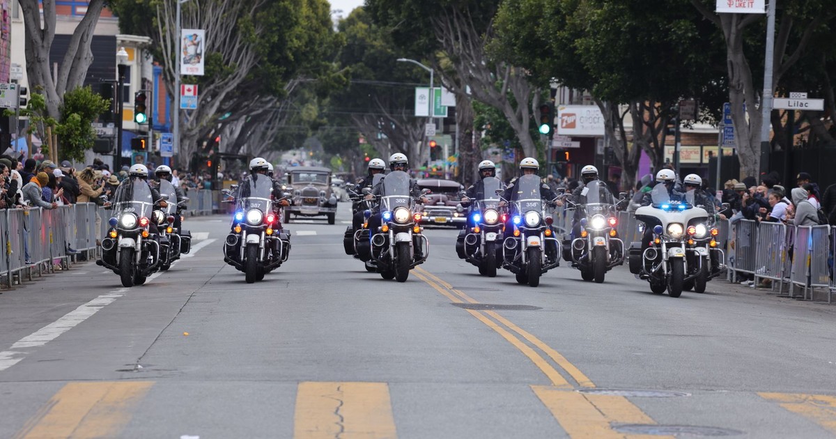 A police motorcade advances through a city street during a parade, with spectators watching.