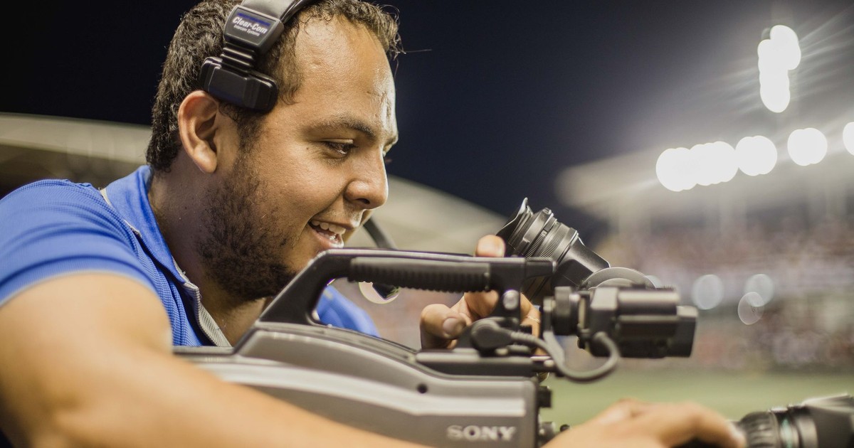 A cameraman with a headset operates a video camera outdoors at night, focusing on capturing footage.