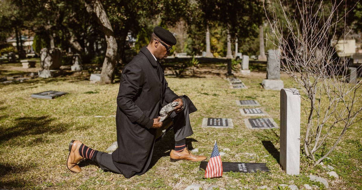 A man kneeling at a gravestone in a cemetery with an American flag.