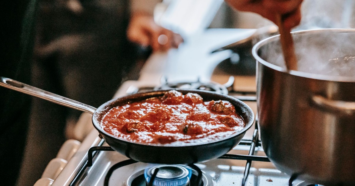 Crop unrecognizable person stirring boiling water in saucepan placed on gas stove near frying pan with appetizing meatballs in tomato sauce