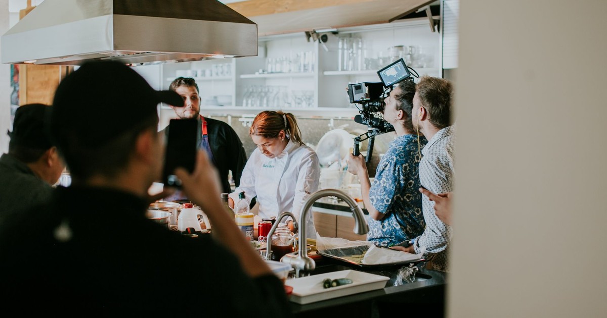 Behind the scenes view of film crew capturing a cooking show in a professional kitchen setting.