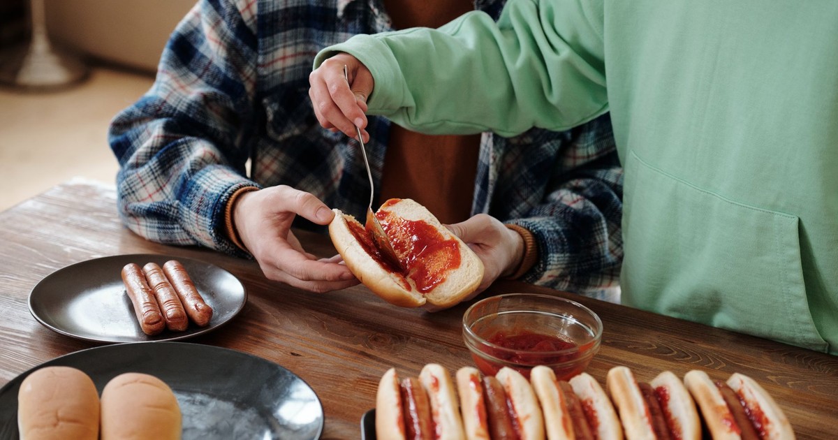 Two people preparing hot dogs with ketchup in an indoor setting, showcasing homemade comfort food.