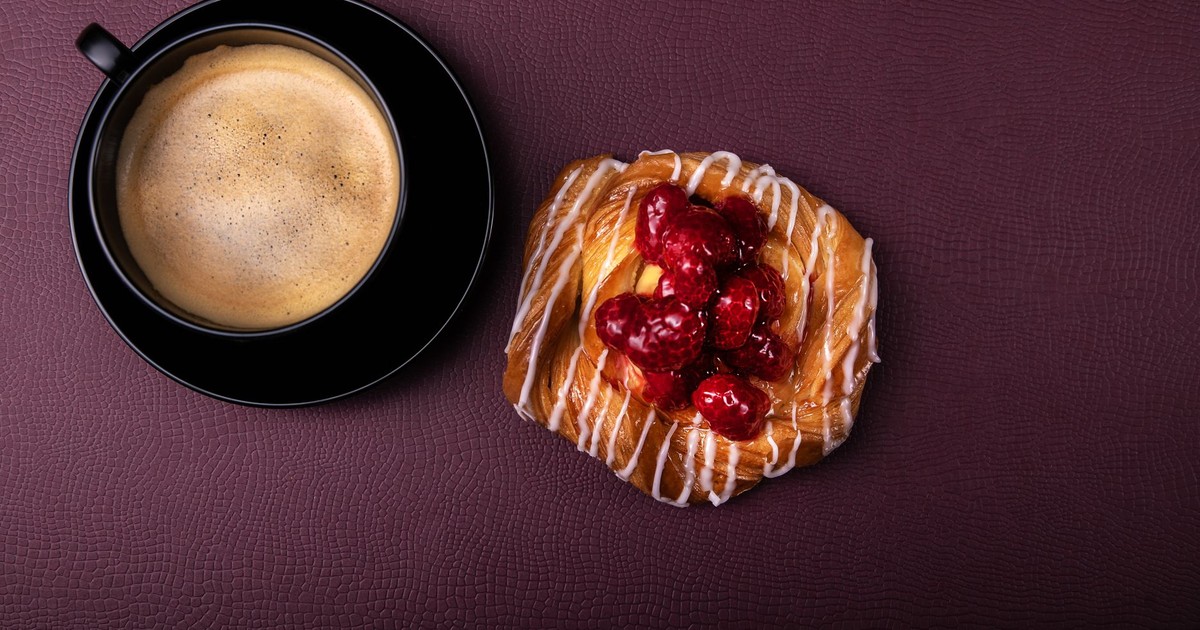 A top-view of a coffee cup and raspberry pastry on a textured surface.