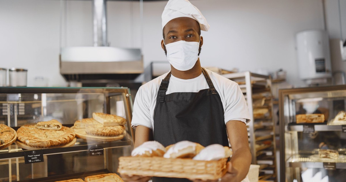 A baker wearing a face mask presenting fresh pastries in a modern bakery setting.