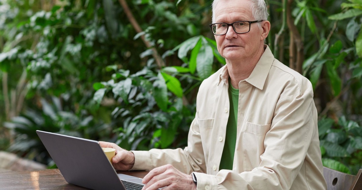 Elderly man using laptop outdoors, surrounded by greenery, remote work concept.