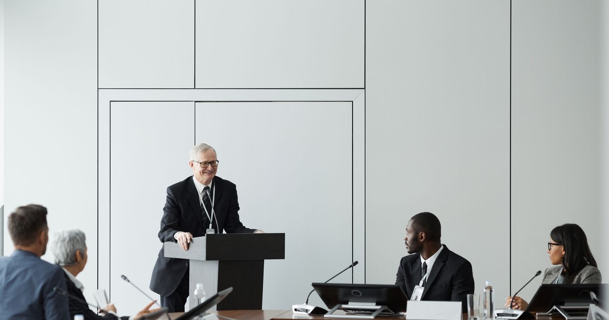 Formal business meeting with a diverse group and speaker at a podium in a conference room.