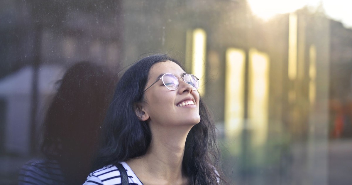 A woman with eyeglasses and striped shirt smiling serenely while leaning against a glass wall outdoors.