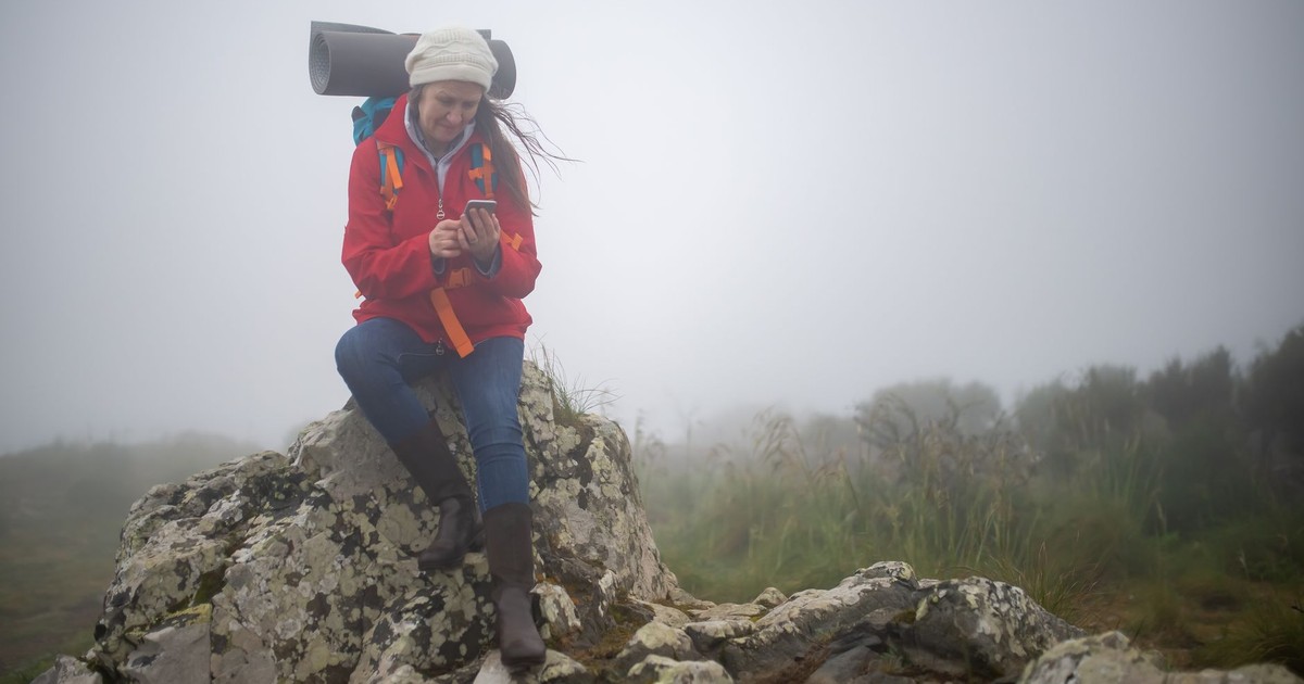 A woman in a red jacket sits on rocks checking her phone amidst a foggy landscape in Portugal.