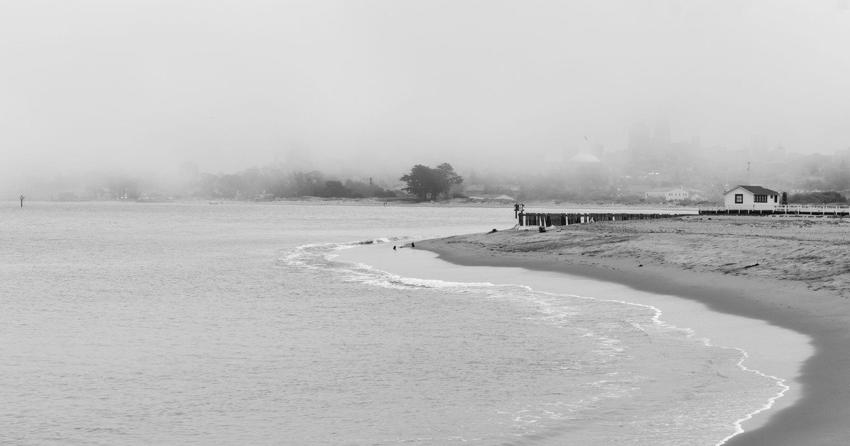 A calm and misty beach scene in black and white, featuring a coastal building and gentle waves.