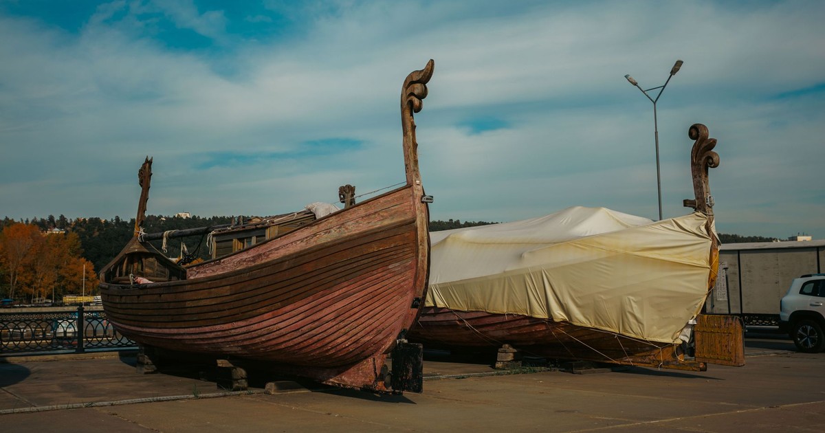 Two wooden Viking ships at a dockside under a clear blue sky, one fully covered.