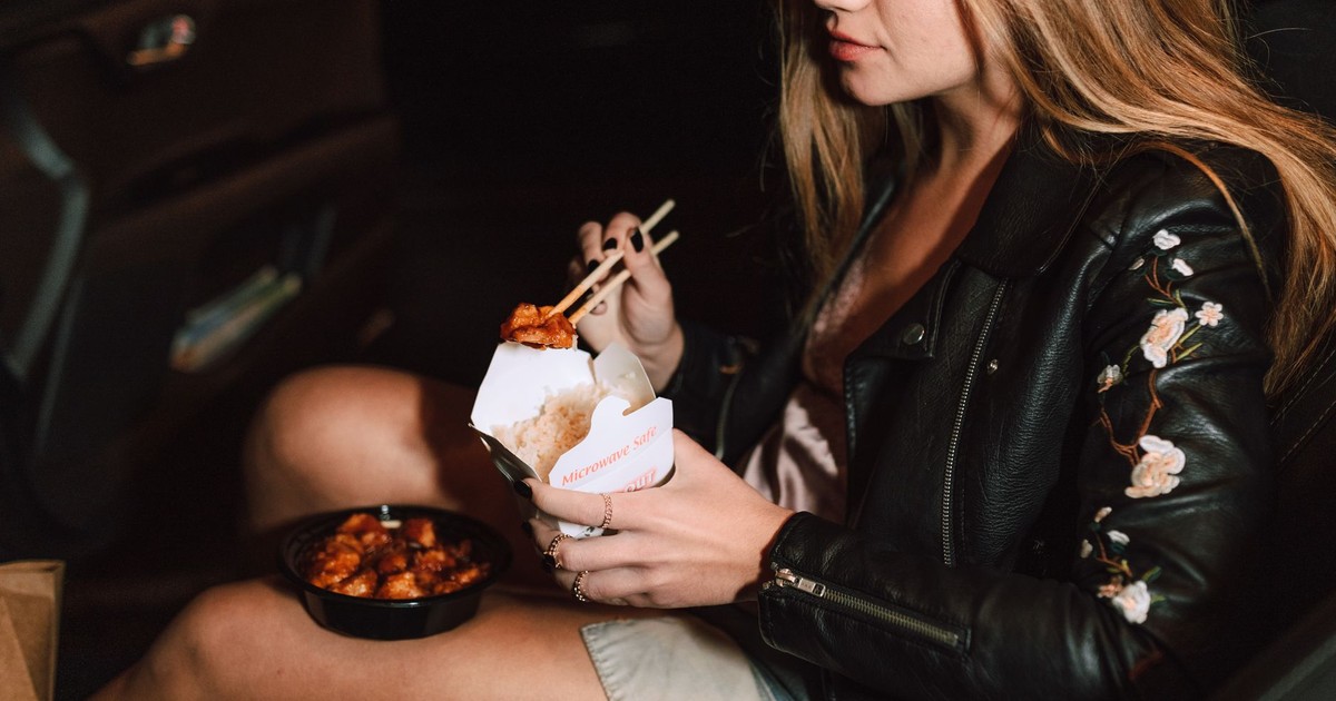 Young woman enjoying Asian takeout food while sitting in a car, wearing a black leather jacket.