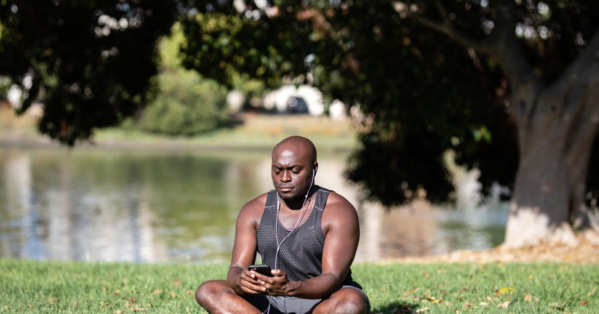 Adult man sitting cross-legged on grass, meditating with smartphone and earphones outdoors.