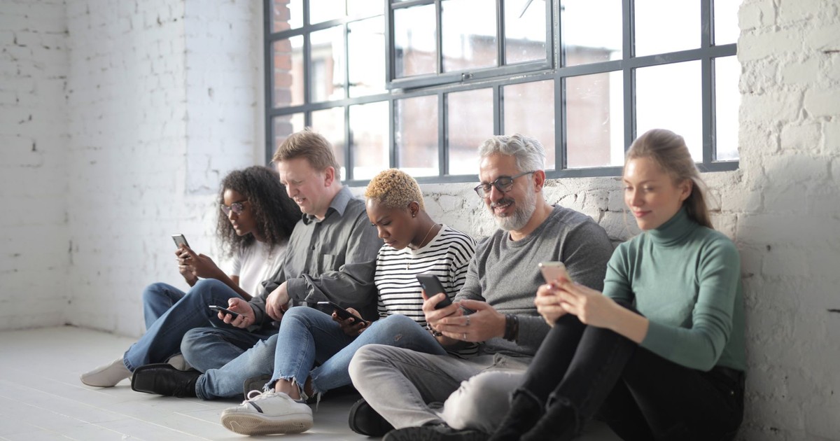 Smiling coworkers of different ages in casual outfit browsing internet on cellphones while sitting with crossed legs near brick wall and fenced window during daytime