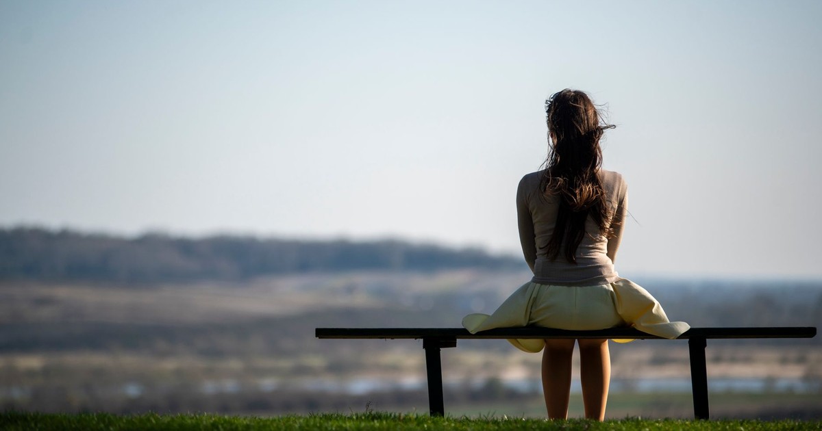 A woman sits on a bench in a field, gazing at the distant horizon under a clear sky.