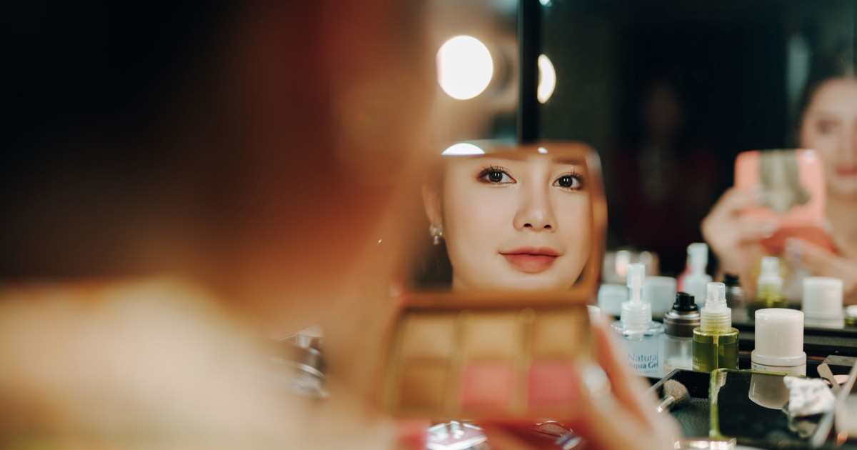 A woman applying makeup, captured in a mirror reflection surrounded by beauty products.
