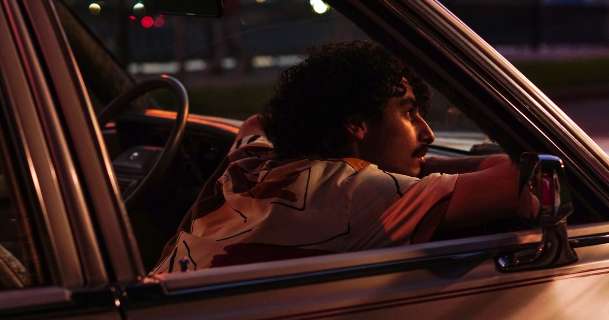 A man with a mustache relaxes in his car at night, captured with moody lighting.