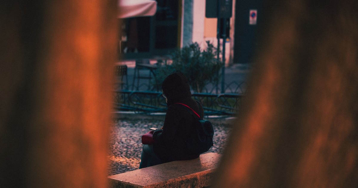 A person wearing a black jacket sits on a bench in an urban area during twilight.