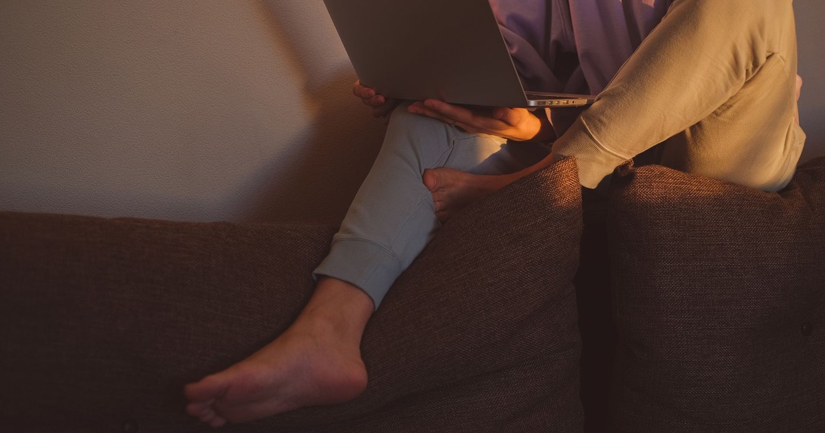 A person sitting barefoot on a couch, using a laptop in warm lighting, indoors.