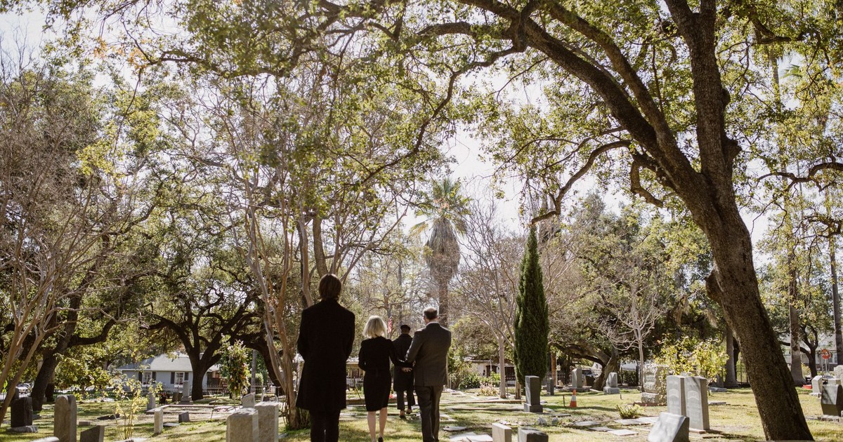 Three people in formal attire walking through a sunlit cemetery.