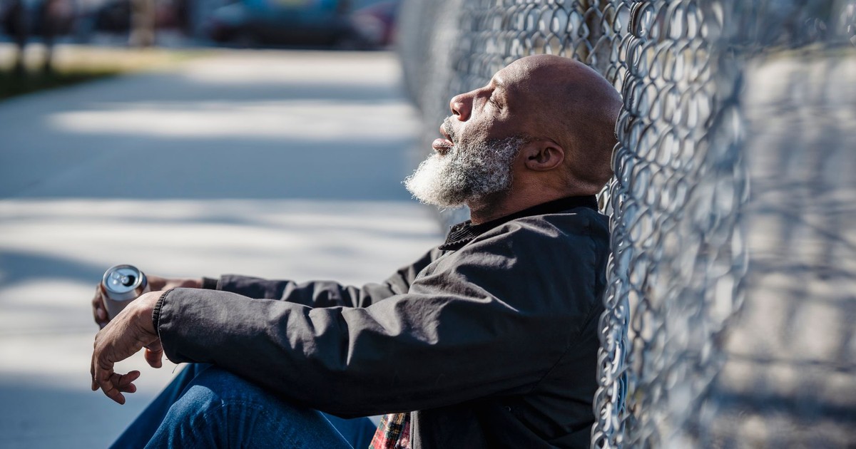 A man with a beard sits against a chain link fence, resting while holding a can, in a city setting.