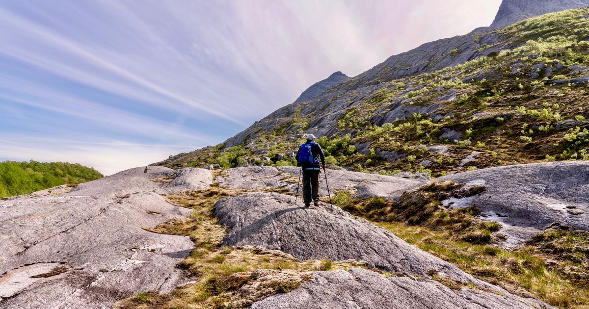 A lone hiker with a blue backpack trekking across a rocky mountain path surrounded by lush spring scenery.