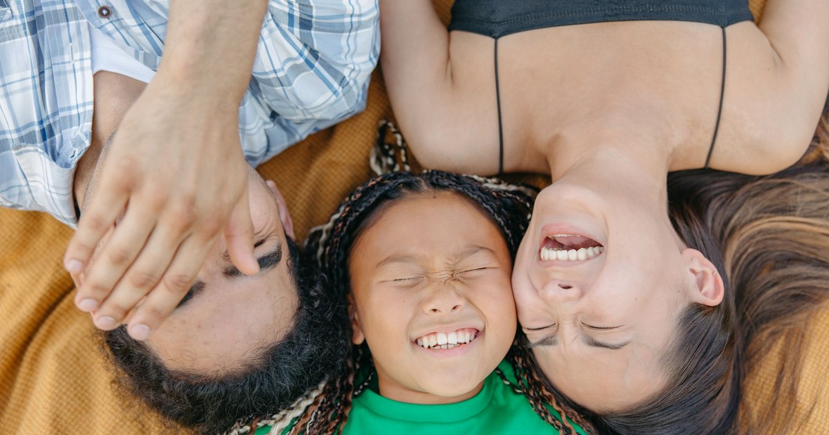 A joyful family bonding moment with parents and daughter laughing while lying on a blanket outdoors.