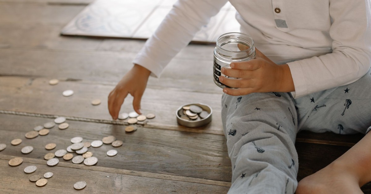 A young child collects coins into a jar on a wooden floor, symbolizing savings.