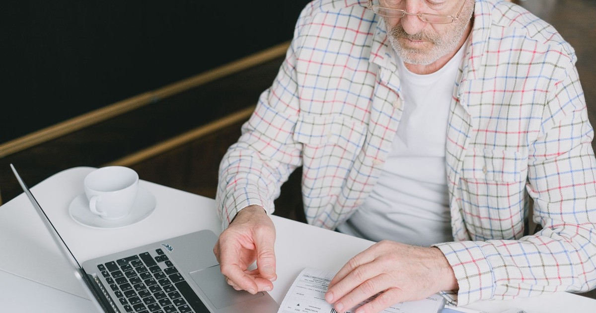 Elderly man with eyeglasses reviewing documents at a laptop. Indoor setting with natural light.