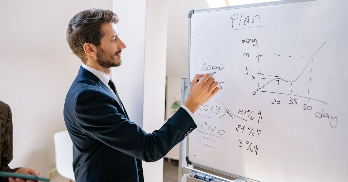 A businessman in a suit writes financial data on a whiteboard during an office planning session.