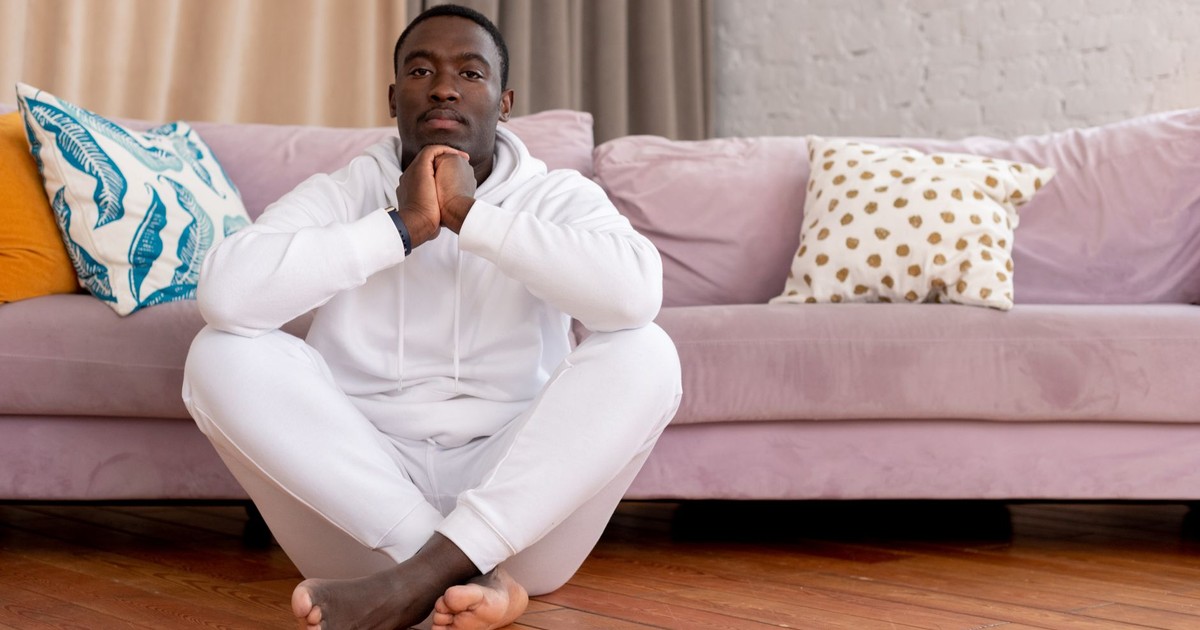 Pensive African American man in light clothes sitting with crossed legs on floor and leaning on pink sofa with pillows in living room in daytime