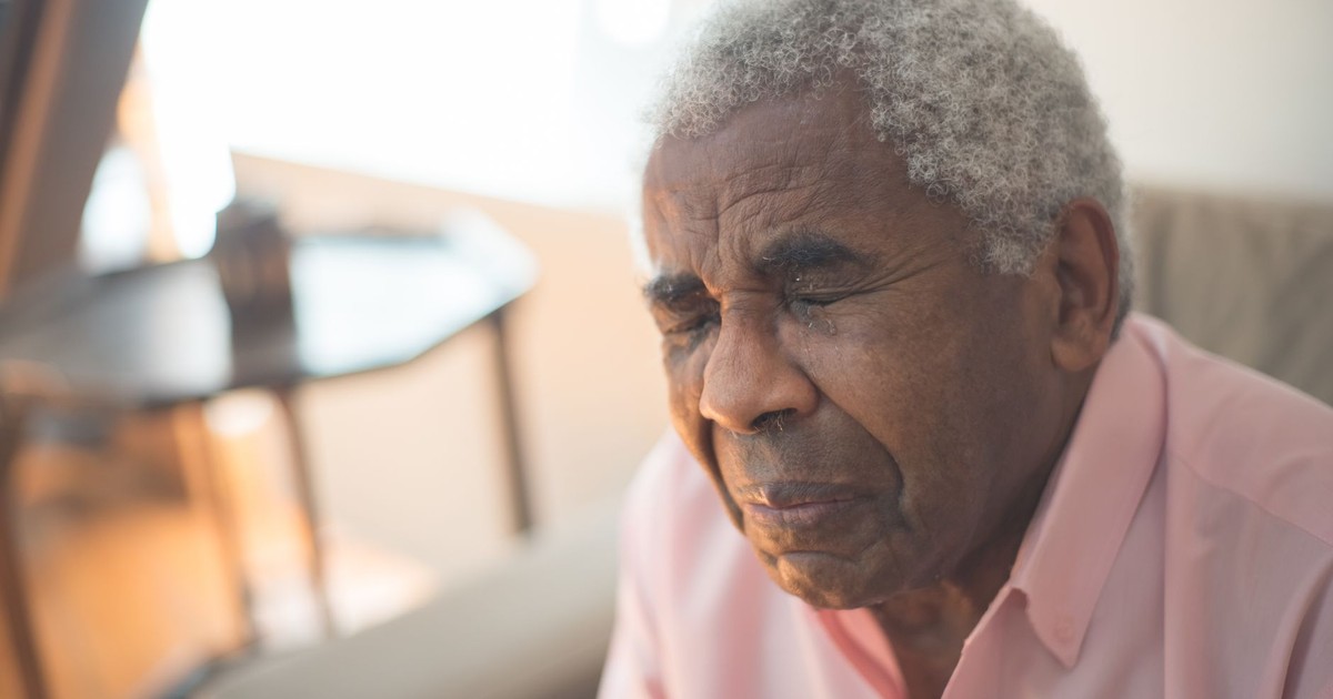 An elderly man with gray hair expresses deep emotion, sitting indoors in a moment of grief.