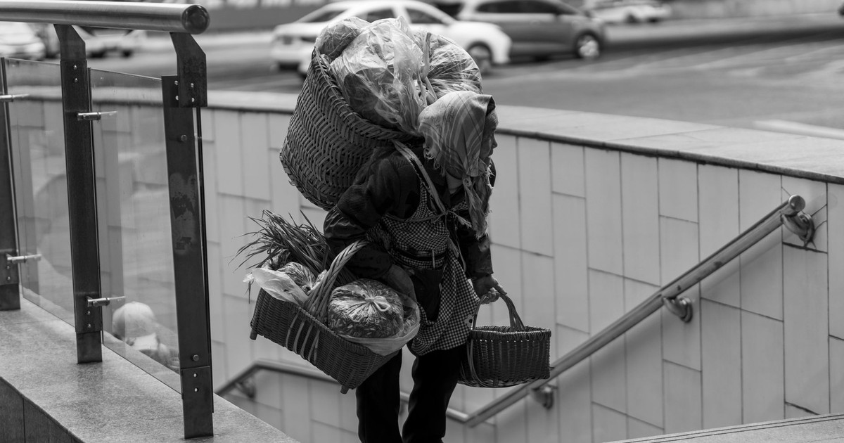A black-and-white photo of a woman carrying multiple baskets up stairs in an urban setting.