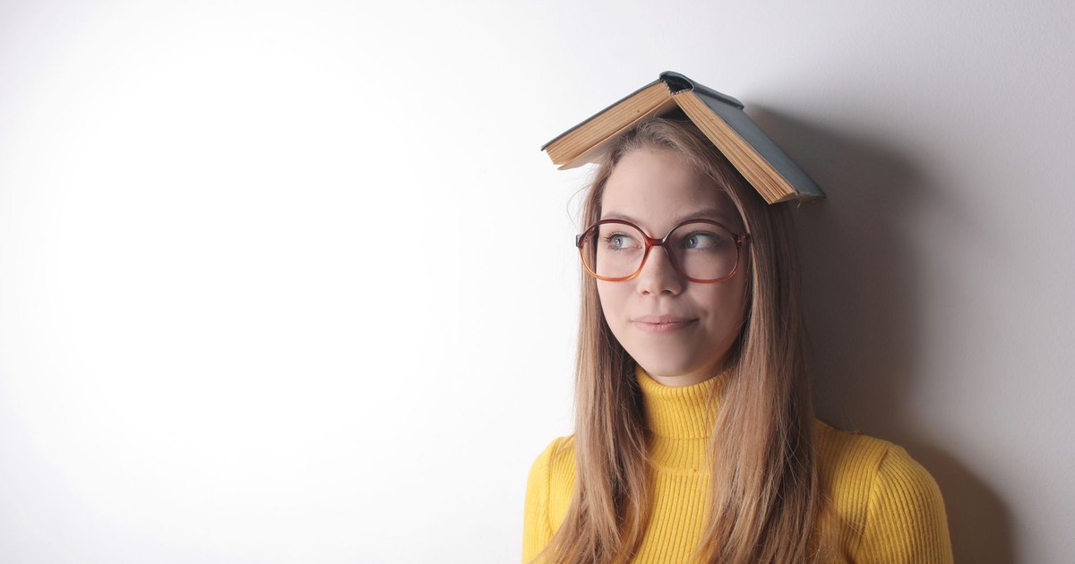 A woman wearing glasses with a book on her head, playfully posing in a yellow sweater.