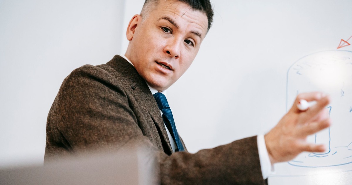 A professional lecturer in formal attire explaining a concept on a whiteboard during an online class session.