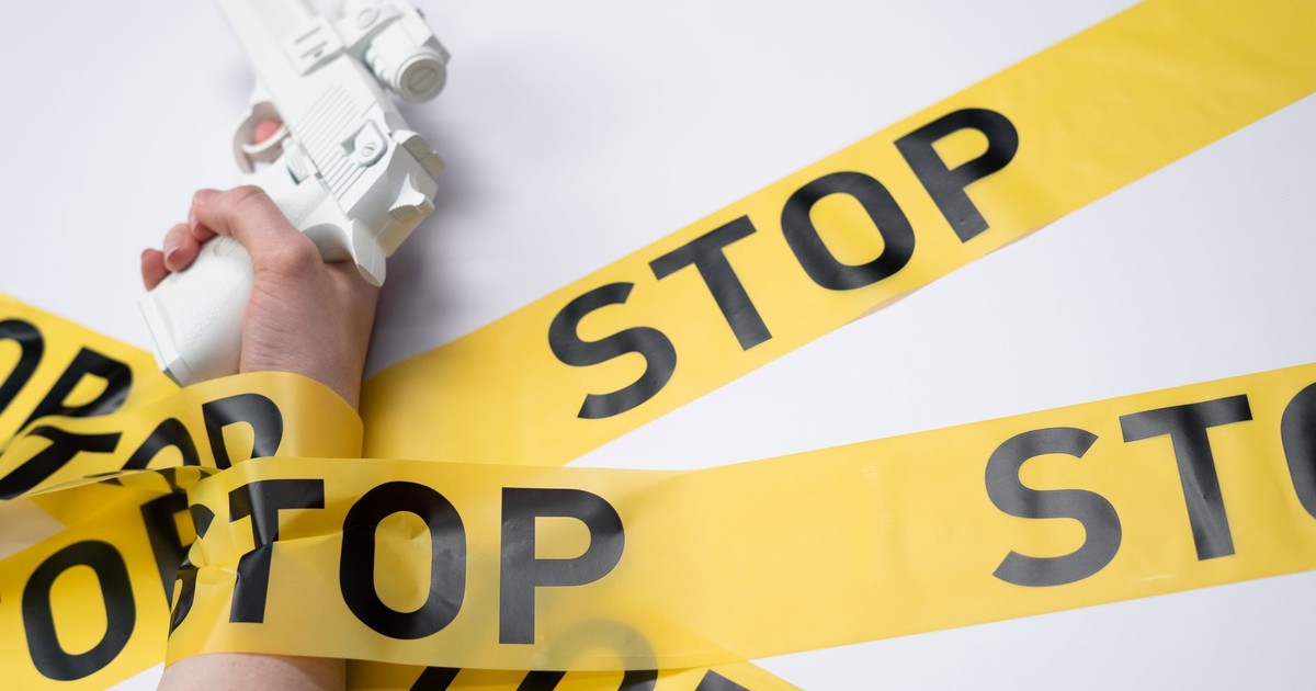 A white toy gun held by a hand wrapped in yellow stop tape on a white background, conveying a message of caution.