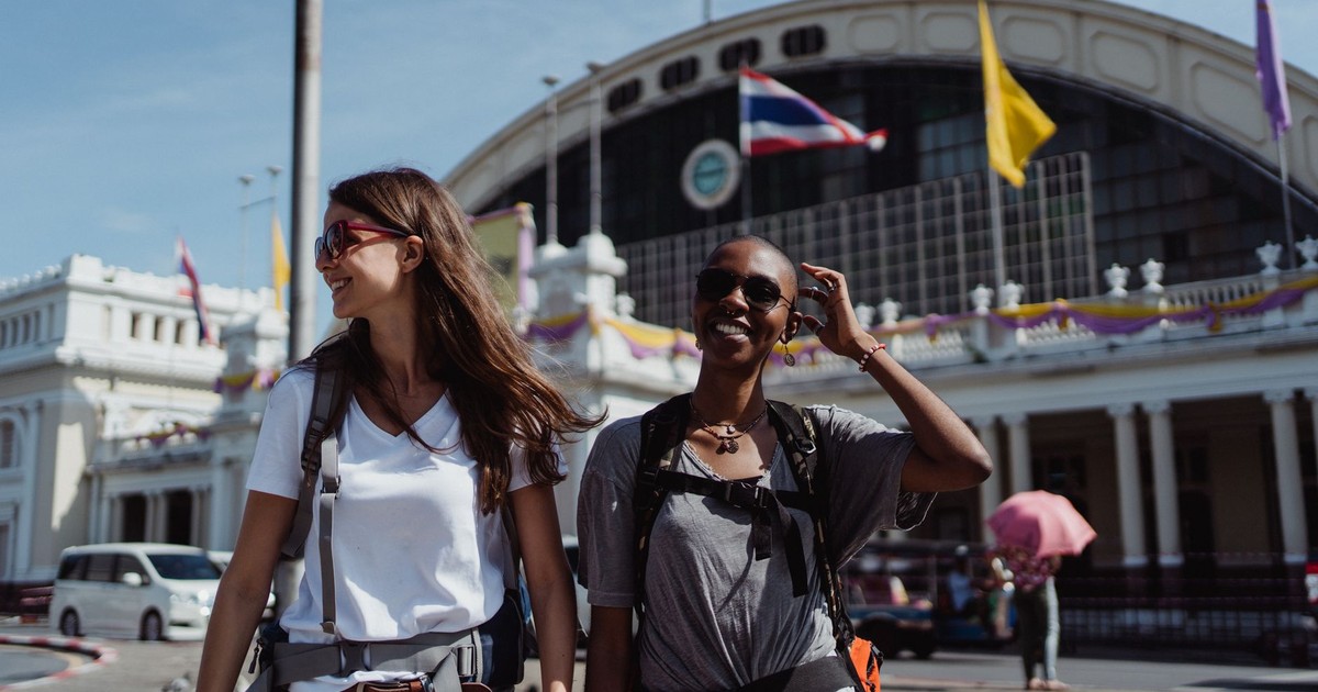 Two women walking and smiling outside in a city setting with a prominent train station in the background.