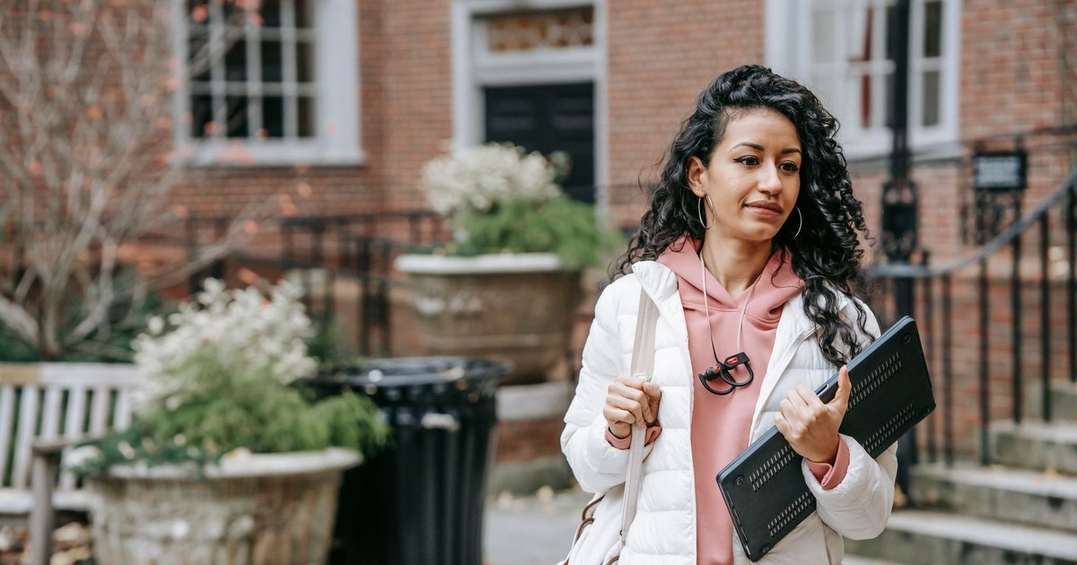 Young ethnic female with long curly hair and netbook in casual trendy outfit on blurred background of brick building