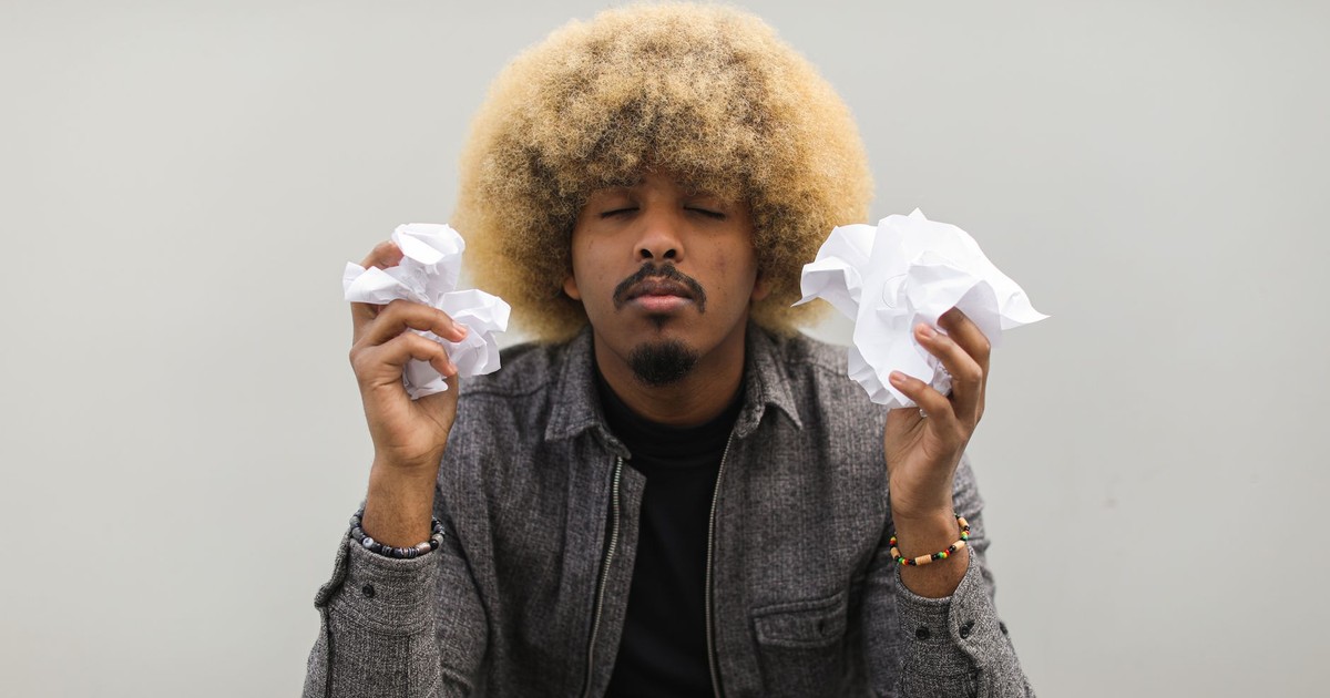 Man with afro hair and beard holding crumpled papers, eyes closed in thoughtful expression.