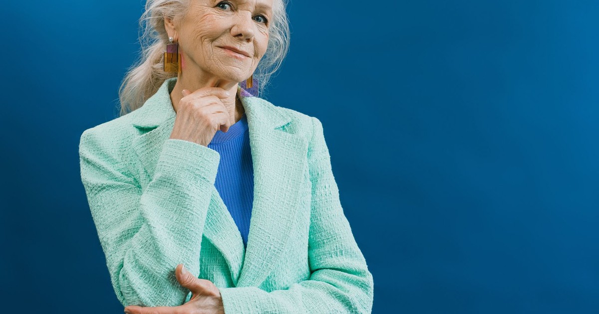 Elderly smiling gray haired female wearing mint blazer and earnings standing against blue background touching chin and looking at camera