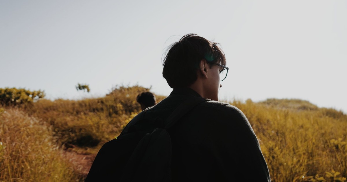 A young man wearing glasses and a backpack hikes a sunlit grassy trail.