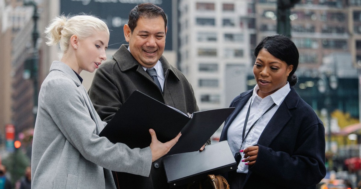 Young well dressed woman sharing folder with multiracial colleagues while talking in town in daytime