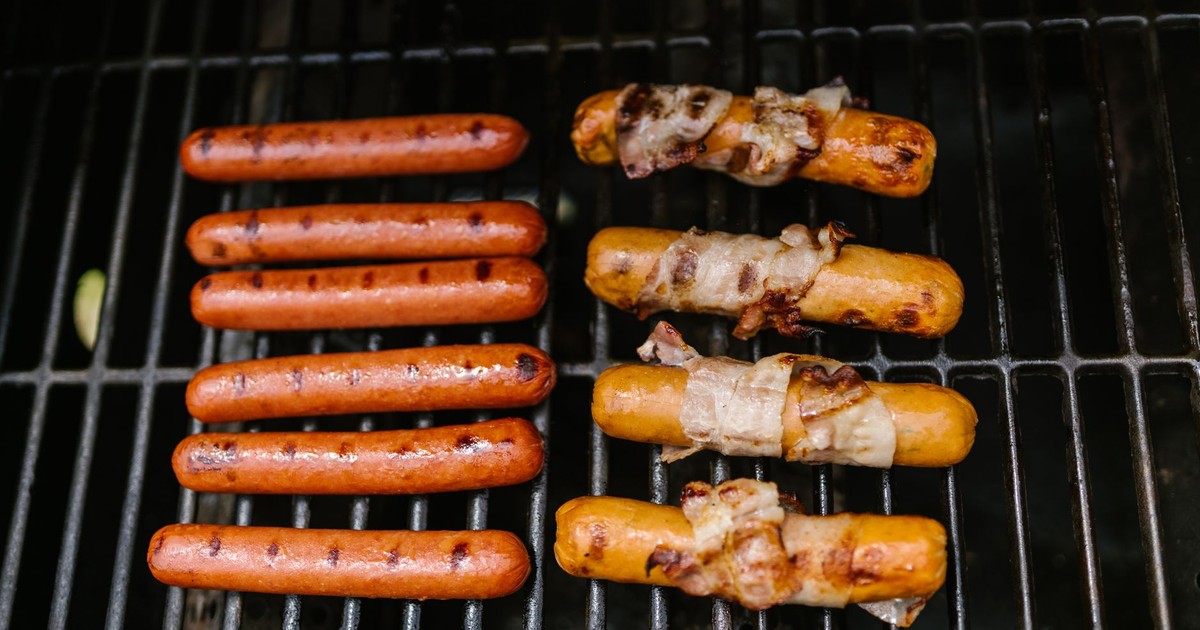 Grilled hot dogs and sausages wrapped in bacon cooking on an outdoor barbecue.
