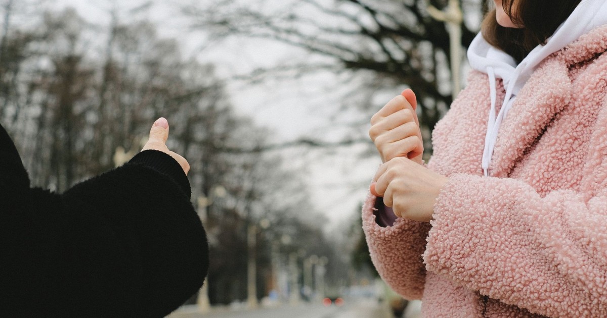 Crop unrecognizable female in warm coat using sign language standing on blurred street and communicating with faceless friend