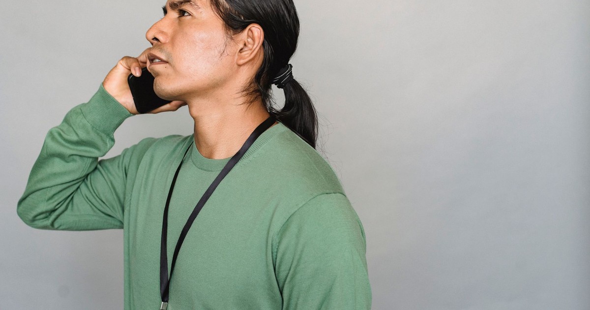 Side view of Native American male worker with ponytail and name tag speaking on cellphone while standing on gray background in studio